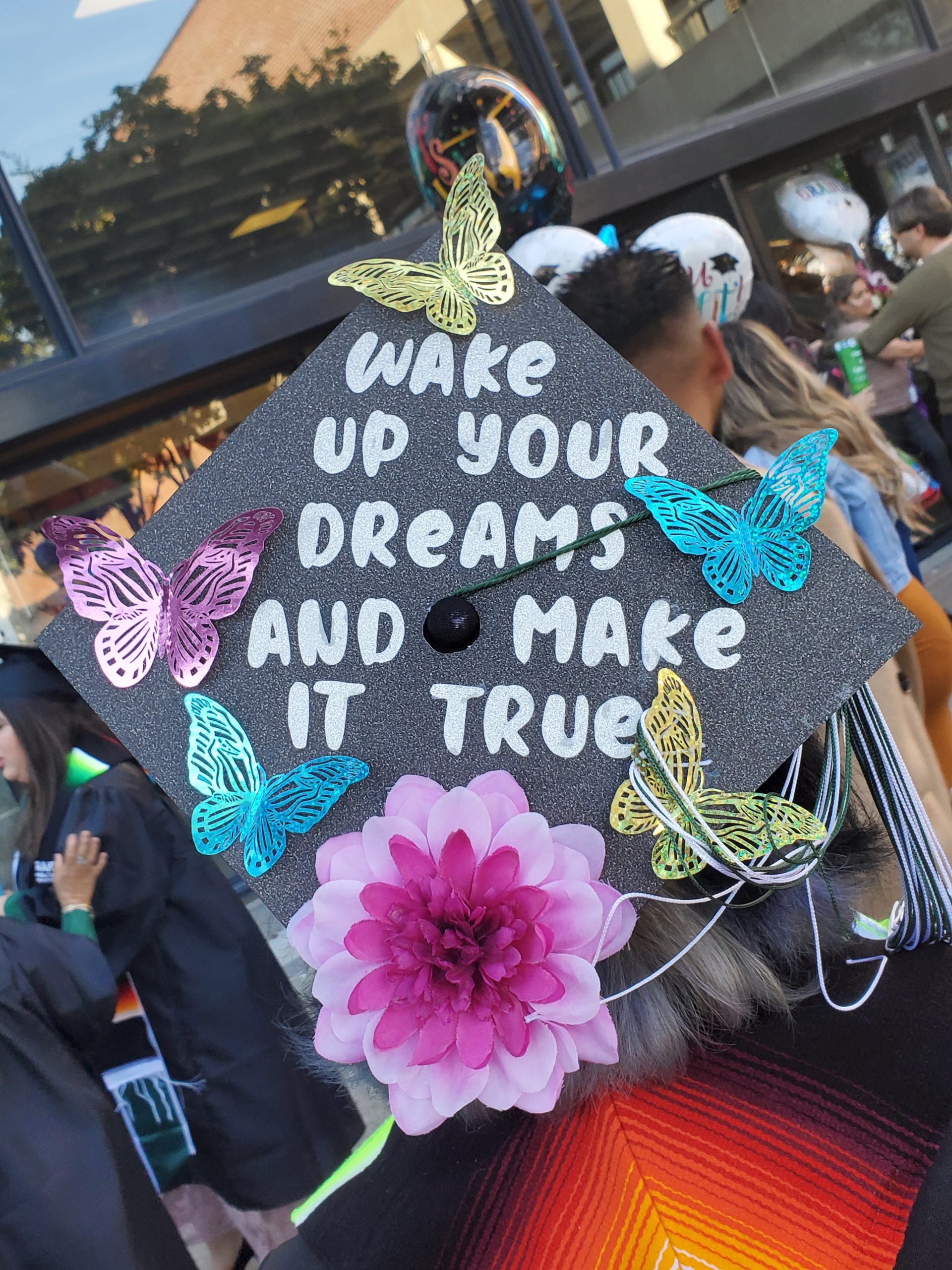 A graduation cap with the words "Wake up your dreams and make it true" glued on the top, along with paper butterflies and a flower